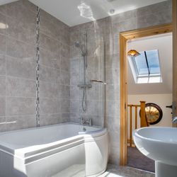 A modern bathroom in Teifi Cottage with a curved bathtub, overhead shower, and sleek grey tiles. A skylight lets in natural light, and an open door leads to the landing.