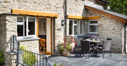 The outdoor patio of Teifi Cottage with a black rattan dining set, vibrant flowers, and stone walls. A door opens to a cosy interior, inviting relaxation in the sunshine.