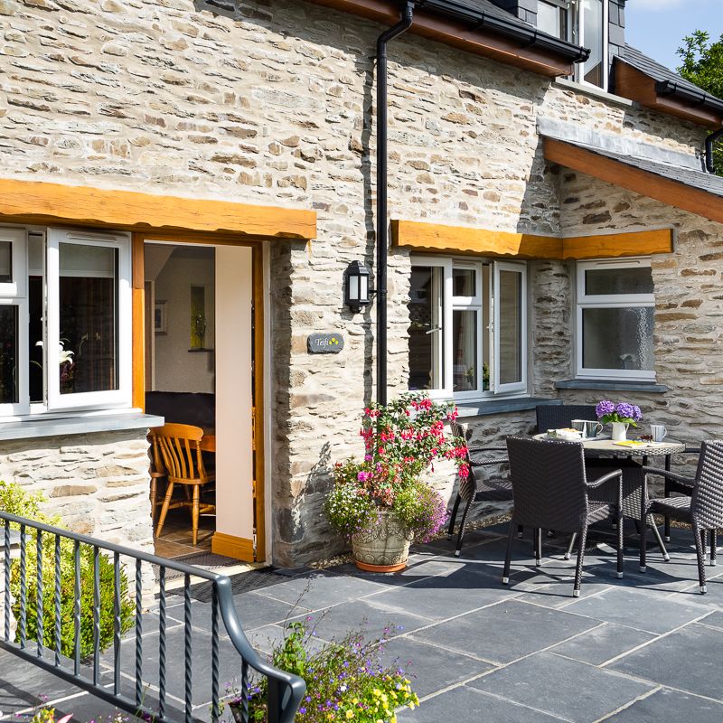 The outdoor patio of Teifi Cottage with a black rattan dining set, vibrant flowers, and stone walls. A door opens to a cosy interior, inviting relaxation in the sunshine.