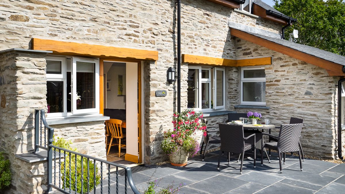 The outdoor patio of Teifi Cottage with a black rattan dining set, vibrant flowers, and stone walls. A door opens to a cosy interior, inviting relaxation in the sunshine.