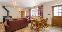 A warm living and dining area in Teifi Cottage with brown leather sofas, a wood-burning stove, a wooden dining table set for a meal, and plaid curtains framing the windows.