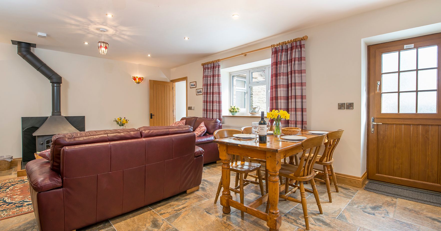 A warm living and dining area in Teifi Cottage with brown leather sofas, a wood-burning stove, a wooden dining table set for a meal, and plaid curtains framing the windows.