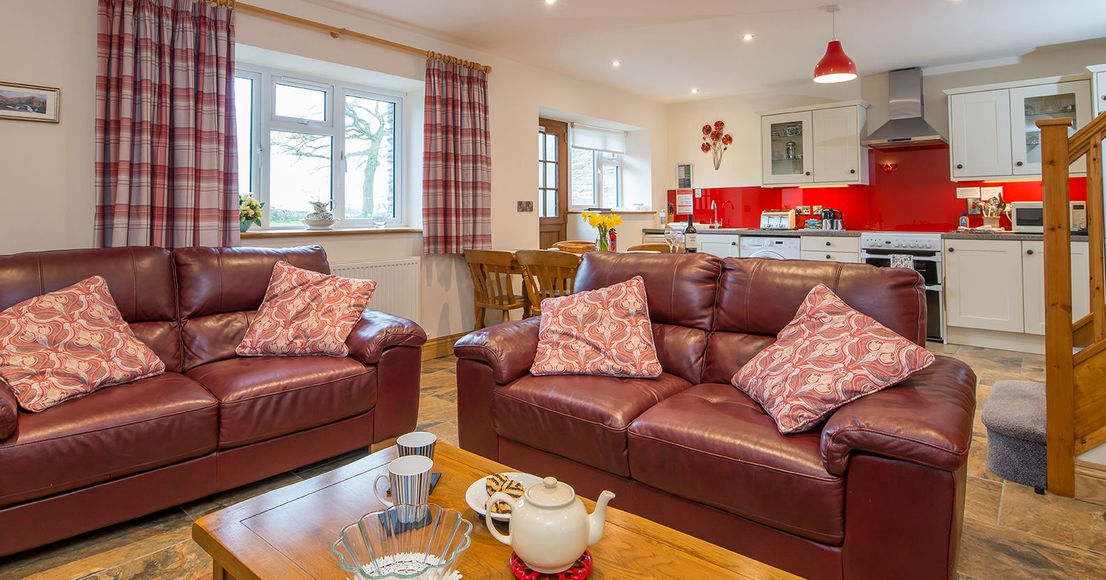 A cosy open-plan living area in Teifi Cottage with brown leather sofas, a wooden coffee table set for tea, and a modern kitchen with red accents in the background.
