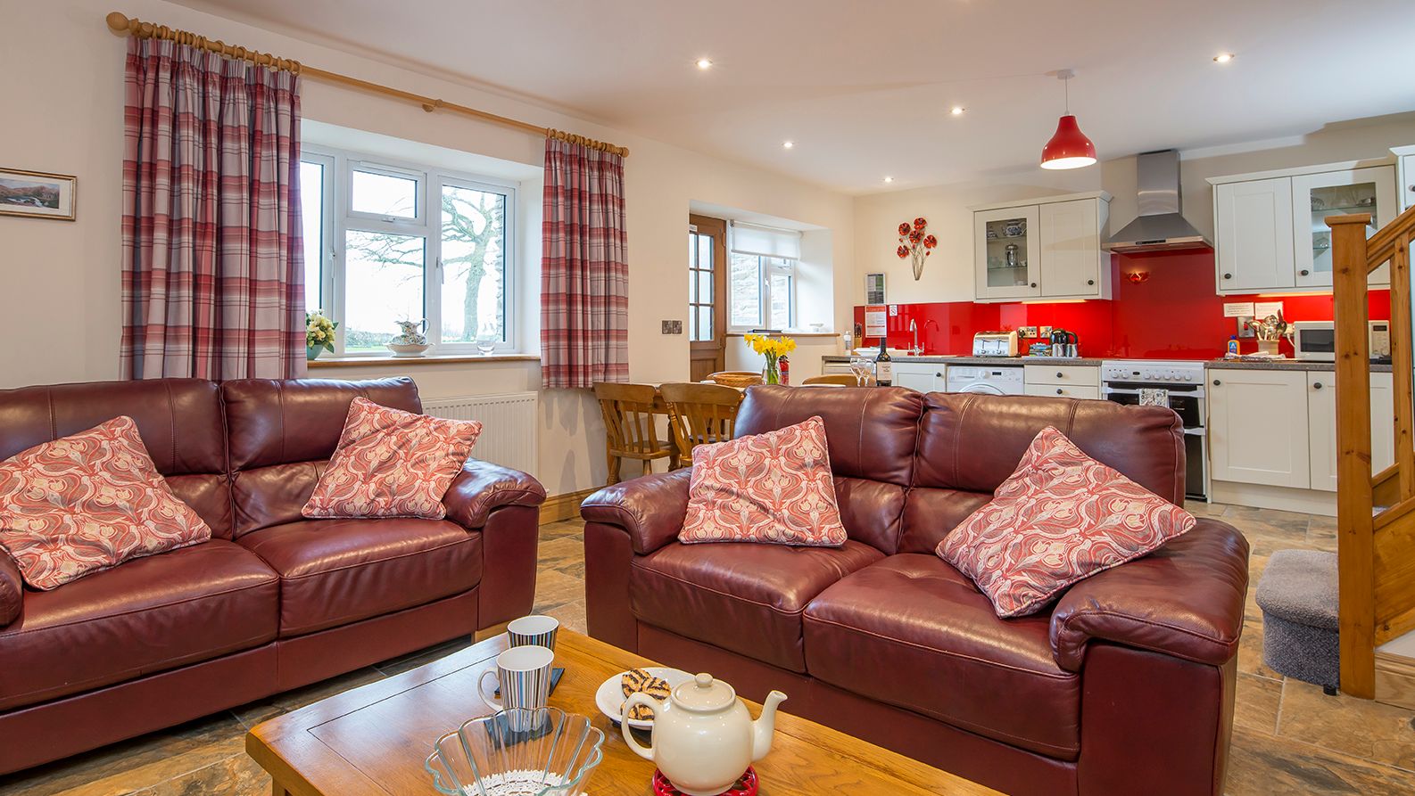 A cosy open-plan living area in Teifi Cottage with brown leather sofas, a wooden coffee table set for tea, and a modern kitchen with red accents in the background.