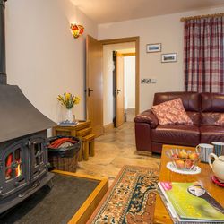 A cosy living room in Teifi Cottage with a wood-burning stove, brown leather sofa, plaid curtains, and a wooden coffee table set with tea, apples, and magazines.