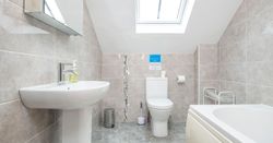 A modern bathroom in Teifi Cottage with a sloped ceiling, skylight, white bathtub, sink, and toilet. Light grey tiled walls and flooring create a bright, clean space.