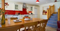 A spacious kitchen and dining area in Teifi Cottage, featuring a wooden table set for a meal, a modern kitchen with red accents, and a staircase leading to the upper floor.