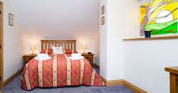 A cosy bedroom in Teifi Cottage with a wooden bed, red and gold quilt, and soft lighting. A stained glass window adds charm, complementing the warm and inviting décor.