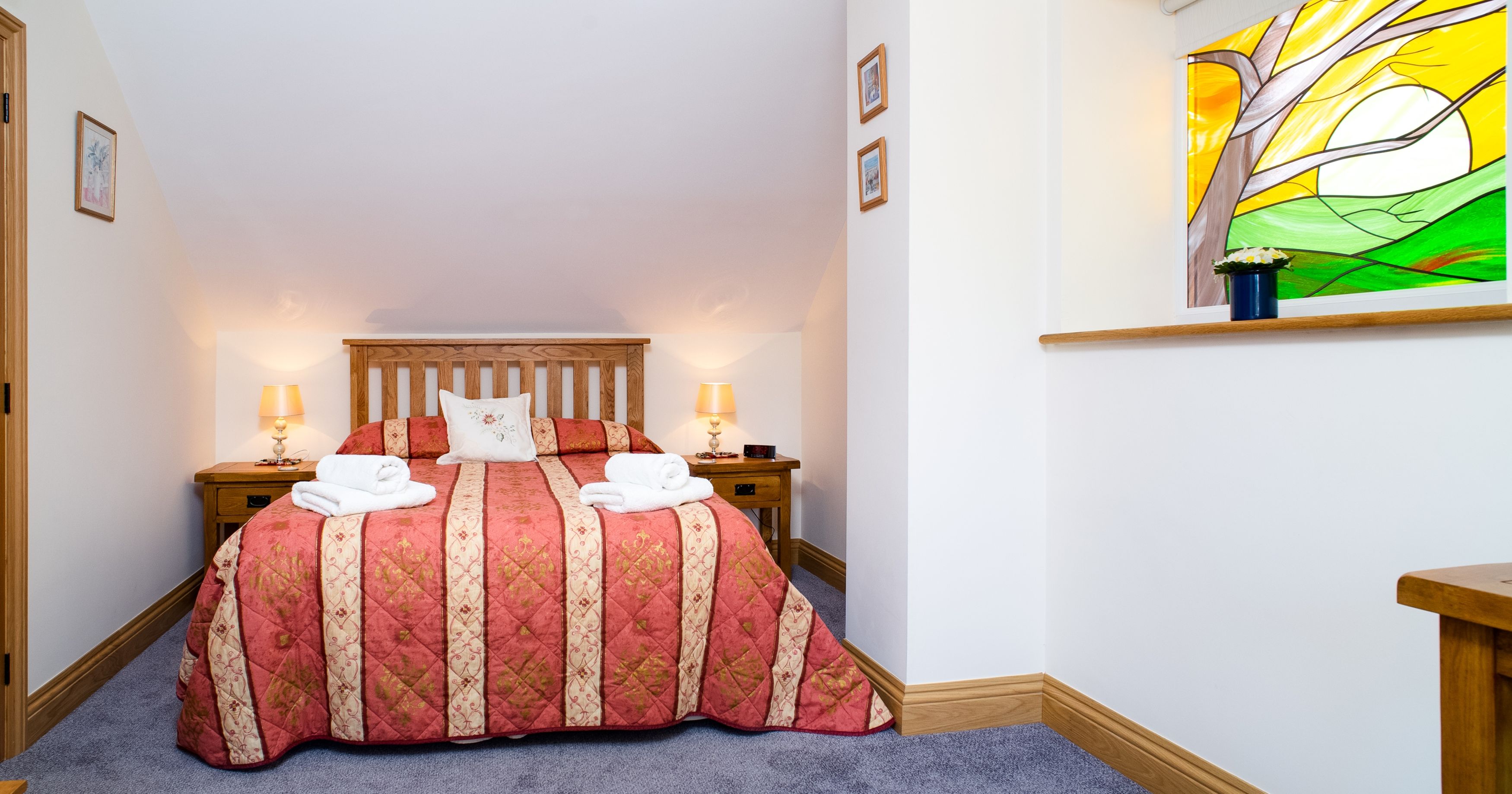 A cosy bedroom in Teifi Cottage with a wooden bed, red and gold quilt, and soft lighting. A stained glass window adds charm, complementing the warm and inviting décor.