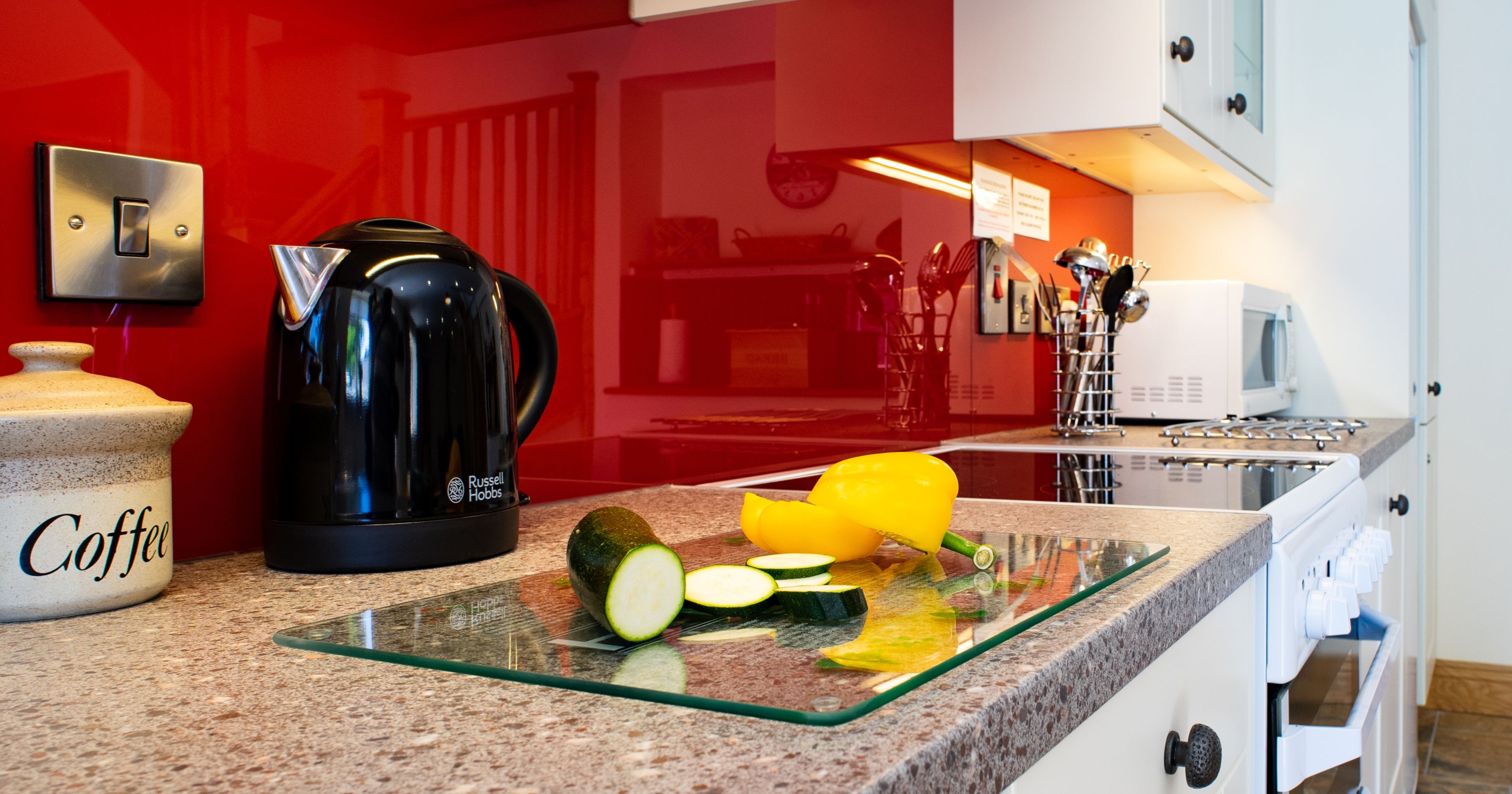 A modern kitchen in Teifi Cottage with a red glass splashback, black kettle, and fresh vegetables on a glass chopping board, creating a vibrant and stylish cooking space.