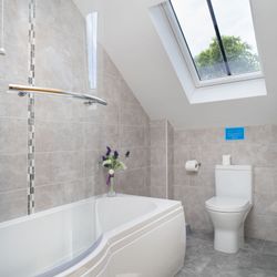 The modern bathroom in Cych Cottage features a curved bathtub with a glass shower screen, a skylight for natural light, and stylish grey tiling for a fresh, airy feel.