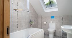 The modern bathroom in Cych Cottage features a curved bathtub with a glass shower screen, a skylight for natural light, and stylish grey tiling for a fresh, airy feel.