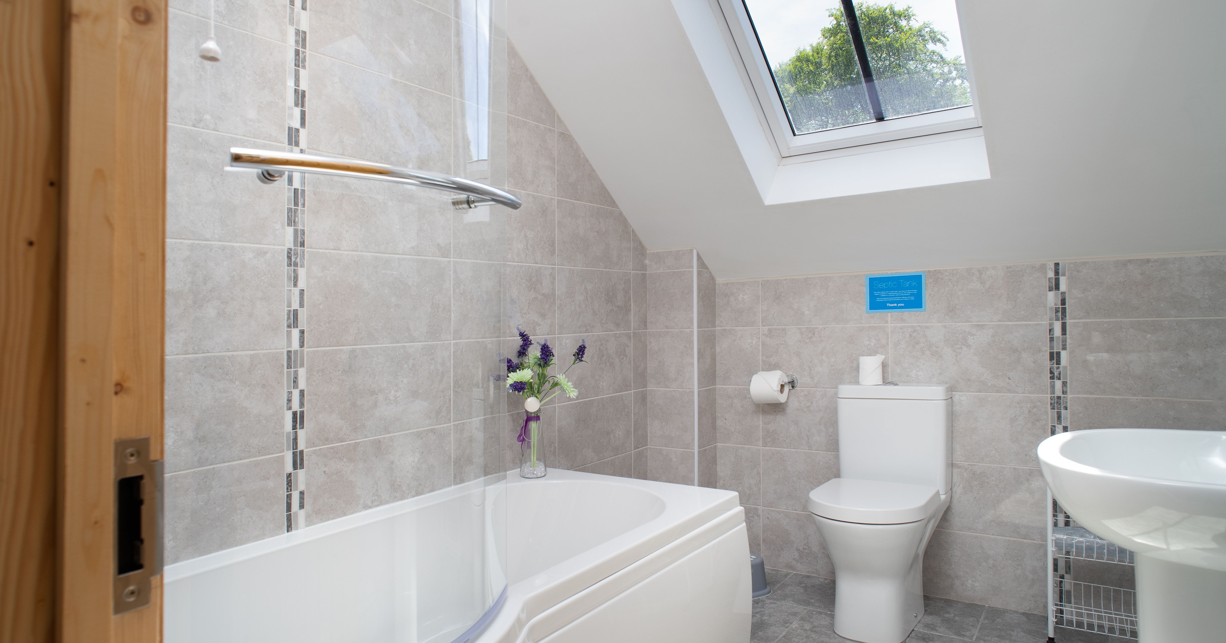 The modern bathroom in Cych Cottage features a curved bathtub with a glass shower screen, a skylight for natural light, and stylish grey tiling for a fresh, airy feel.