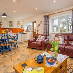 The open-plan living area in Cych Cottage features a cosy kitchen with an orange backsplash, a dining table, and leather sofas with plaid cushions, creating a warm, inviting space.