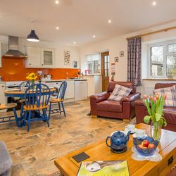 The open-plan living area in Cych Cottage features a cosy kitchen with an orange backsplash, a dining table, and leather sofas with plaid cushions, creating a warm, inviting space.