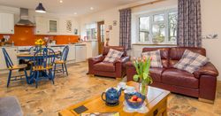 The open-plan living area in Cych Cottage features a cosy kitchen with an orange backsplash, a dining table, and leather sofas with plaid cushions, creating a warm, inviting space.