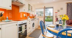 The kitchen in Cych Cottage has cream cabinets, an orange backsplash, and a blue dining set. The open door leads to a patio with garden views, bringing in natural light.