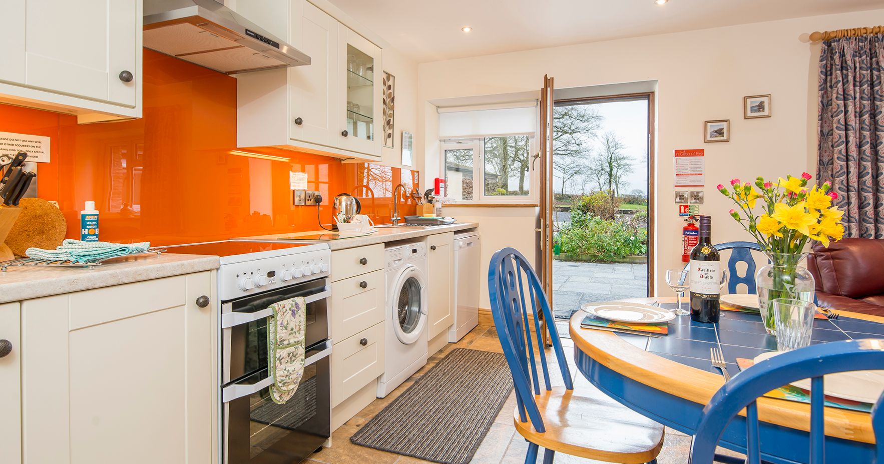 The kitchen in Cych Cottage has cream cabinets, an orange backsplash, and a blue dining set. The open door leads to a patio with garden views, bringing in natural light.