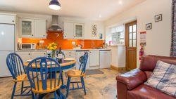 The kitchen in Cych Cottage features cream cabinets, an orange backsplash, and a blue dining set. A cosy living area with a leather sofa sits near the wooden door.