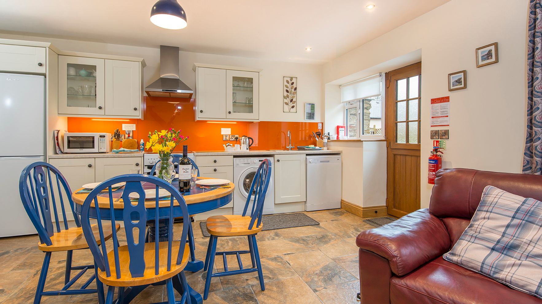 The kitchen in Cych Cottage features cream cabinets, an orange backsplash, and a blue dining set. A cosy living area with a leather sofa sits near the wooden door.