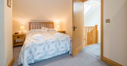 A cozy bedroom in Cych Cottage with a wooden bed, soft lighting, and a quilted bedspread. An open door leads to a bright landing with a wooden staircase and skylight.