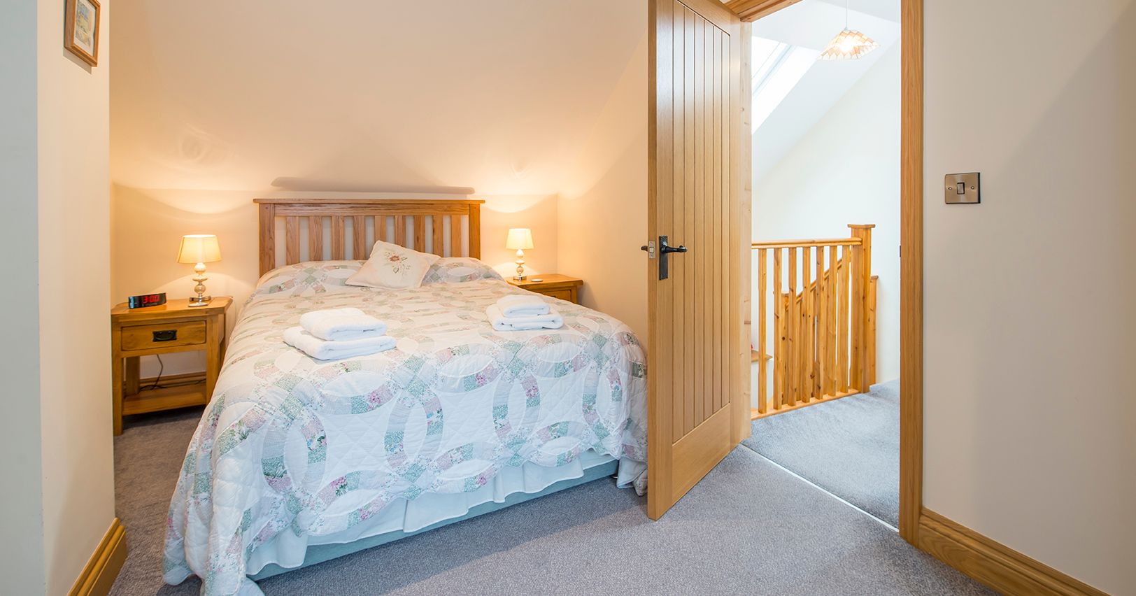 A cozy bedroom in Cych Cottage with a wooden bed, soft lighting, and a quilted bedspread. An open door leads to a bright landing with a wooden staircase and skylight.