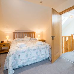 A cozy bedroom in Cych Cottage with a wooden bed, soft lighting, and a quilted bedspread. An open door leads to a bright landing with a wooden staircase and skylight.