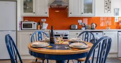 The kitchen in Cych Cottage features cream cabinets, an orange backsplash, and a round wooden table with blue chairs set for a meal with wine, cheese, and grapes.