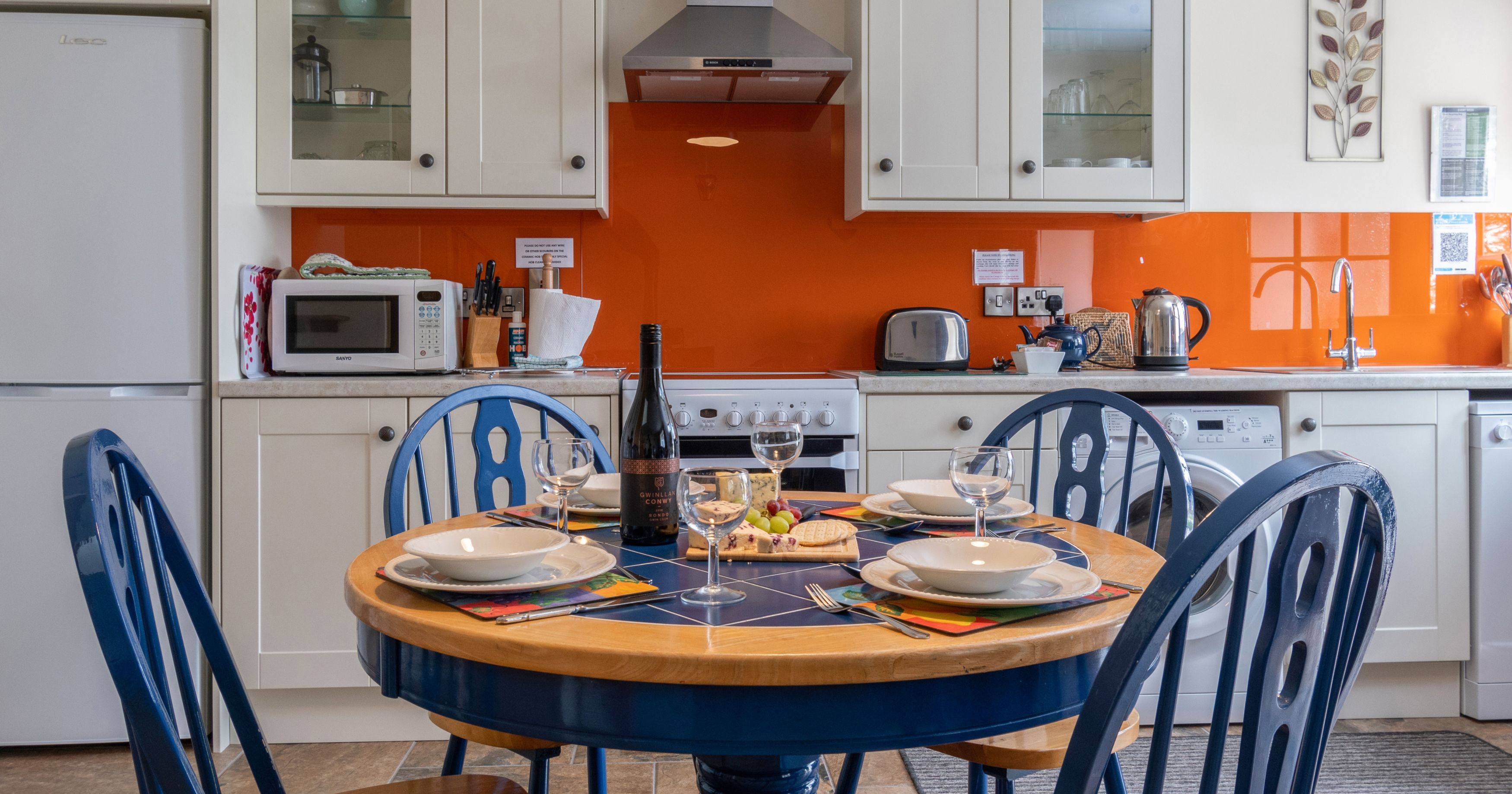 The kitchen in Cych Cottage features cream cabinets, an orange backsplash, and a round wooden table with blue chairs set for a meal with wine, cheese, and grapes.
