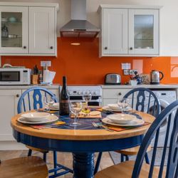 The kitchen in Cych Cottage features cream cabinets, an orange backsplash, and a round wooden table with blue chairs set for a meal with wine, cheese, and grapes.