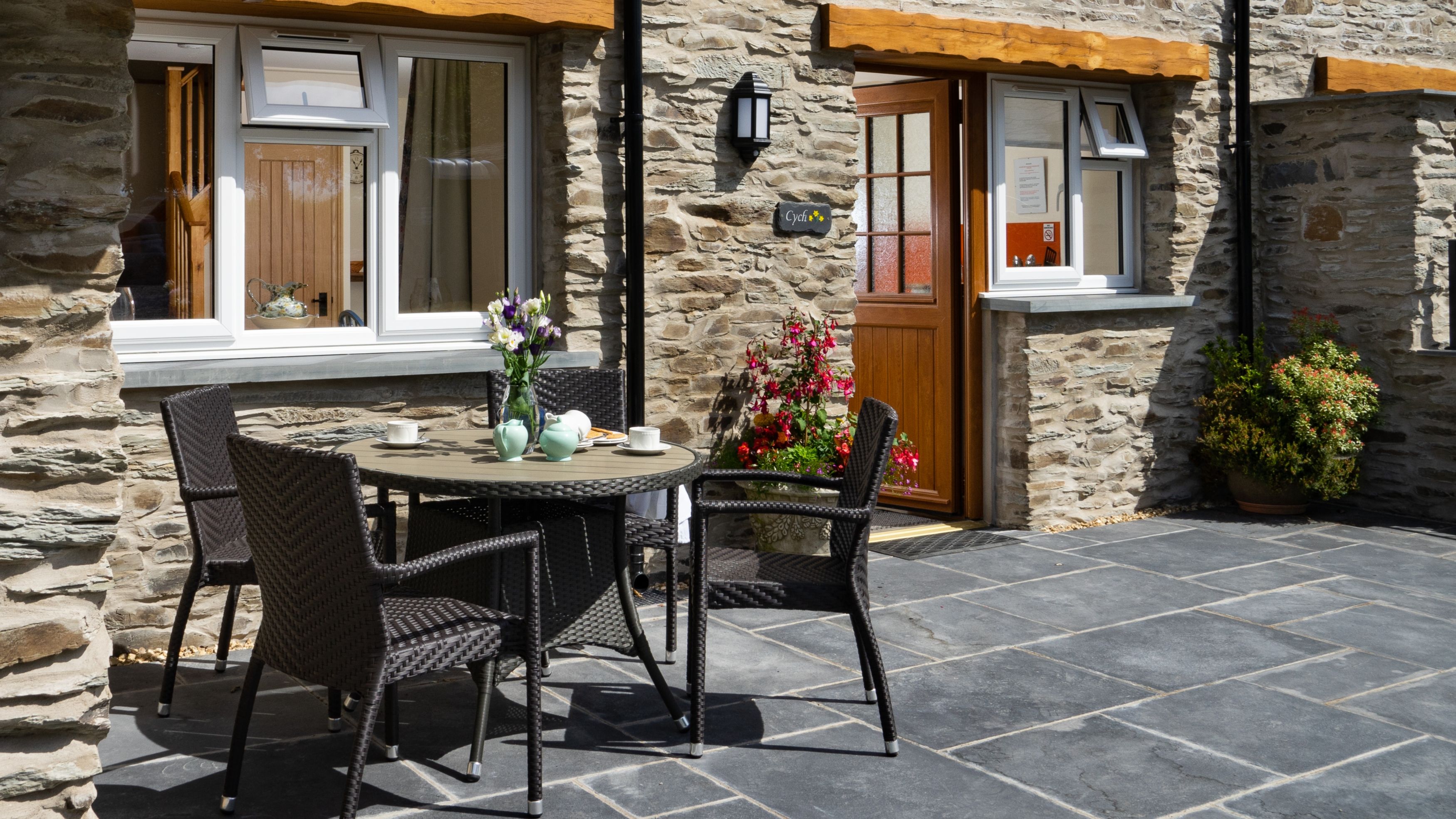 Cych Cottage, a charming stone house with a wooden door, features a sunny patio with a rattan dining set, fresh flowers, and potted plants, creating a welcoming outdoor space.