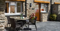 Cych Cottage, a charming stone house with a wooden door, features a sunny patio with a rattan dining set, fresh flowers, and potted plants, creating a welcoming outdoor space.