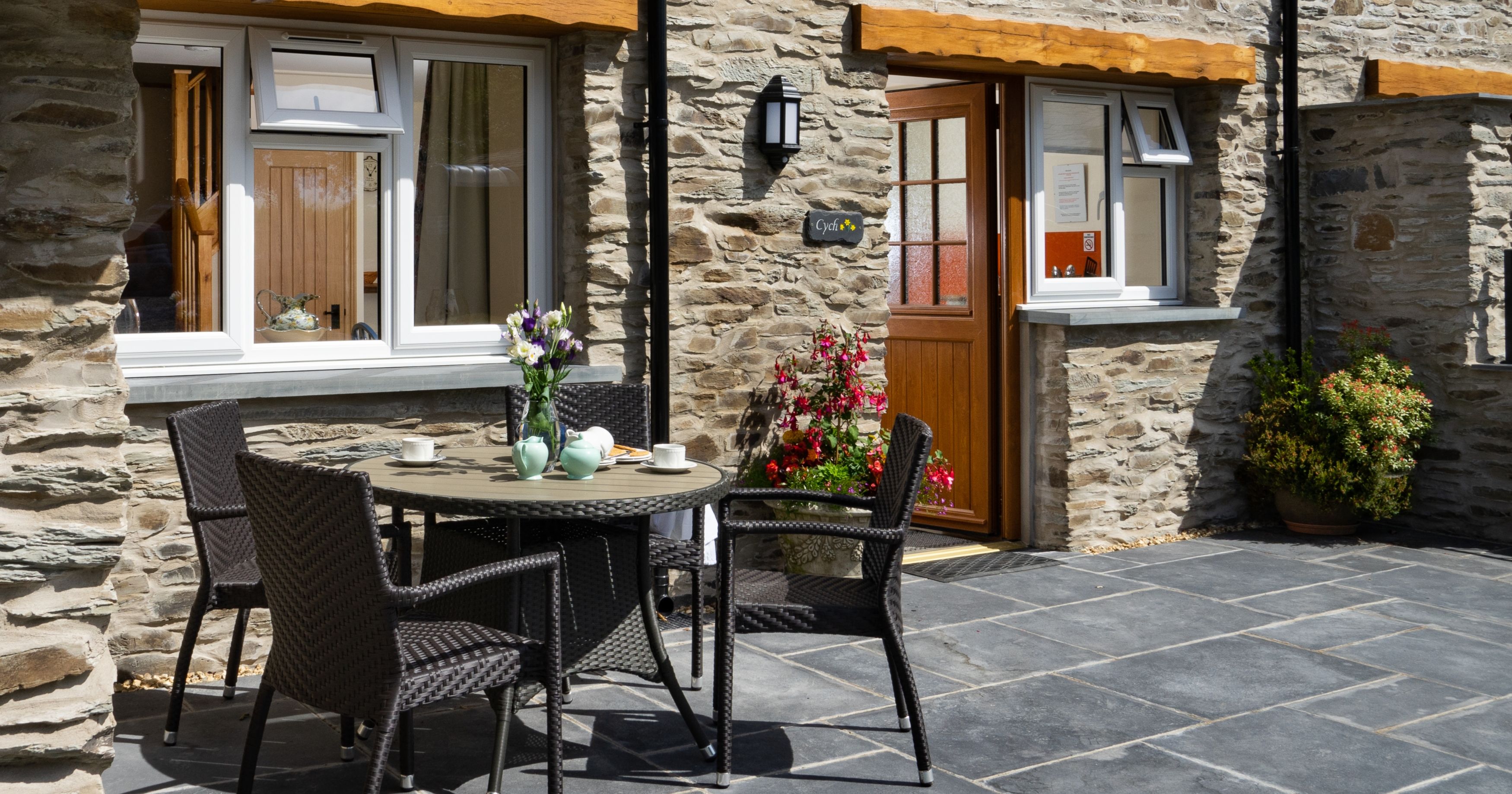 Cych Cottage, a charming stone house with a wooden door, features a sunny patio with a rattan dining set, fresh flowers, and potted plants, creating a welcoming outdoor space.