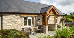 Gwaun Cottage, a stone house with a slate roof and wooden porch, has a cozy patio with seating, surrounded by lush greenery and colourful flowers.
