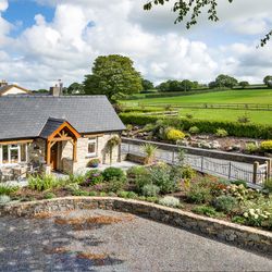 Gwaun Cottage, a charming stone house with a slate roof and wooden porch, is surrounded by lush gardens. A patio with seating overlooks rolling green fields and pastures.