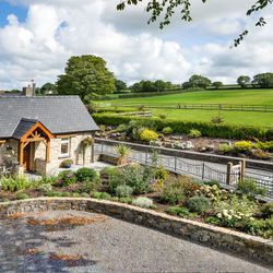 Gwaun Cottage, a charming stone house with a slate roof and wooden porch, is surrounded by lush gardens. A patio with seating overlooks rolling green fields and pastures.