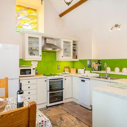 A bright country kitchen with cream cabinets, a green backsplash, and modern appliances. A dining table with wine and flowers sits nearby, beneath a colorful stained-glass window.
