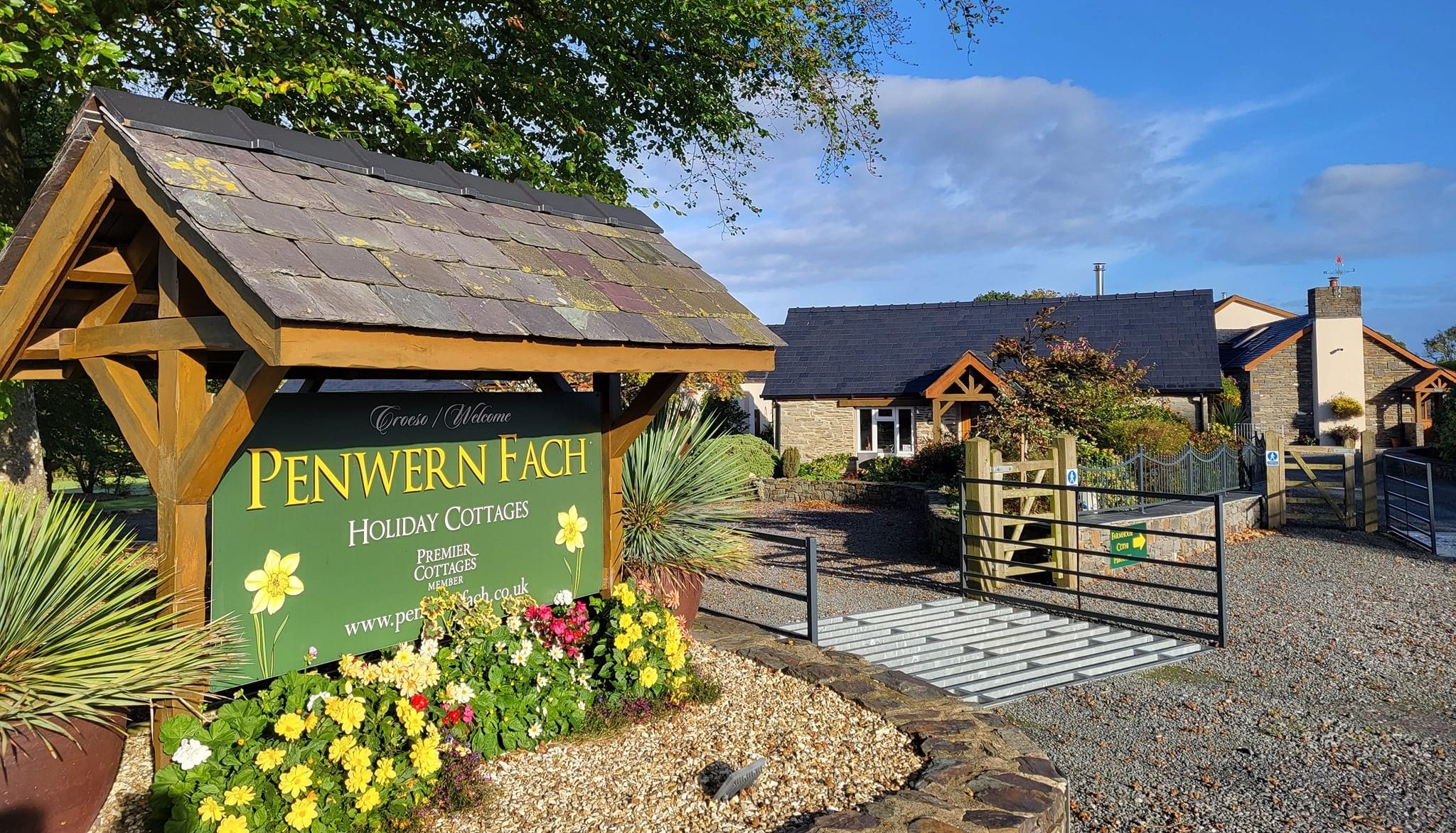 Entrance to Penwern Fach Holiday Cottages, featuring a wooden sign with yellow flowers, a gravel driveway, stone cottages with slate roofs, and a bright blue sky overhead.