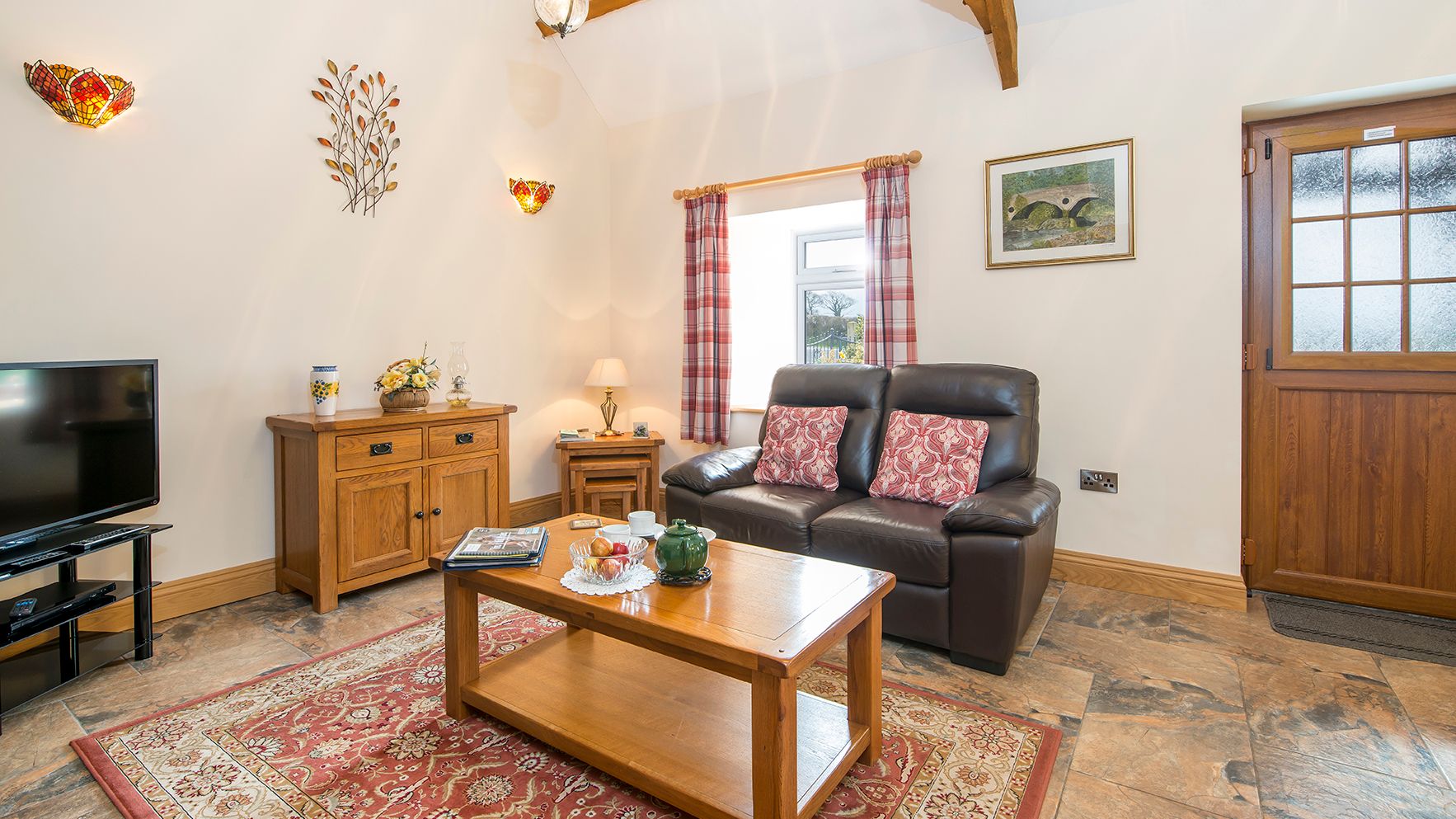 A cozy living room with a leather sofa, wooden furniture, and a red patterned rug. A coffee table is set for tea, with a TV, wall décor, and a wooden door adding charm.