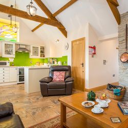 A cozy open-plan living space with a wood-burning stove, a stone feature wall, and a leather armchair. A wooden coffee table is set for tea, with a bright kitchen nearby.