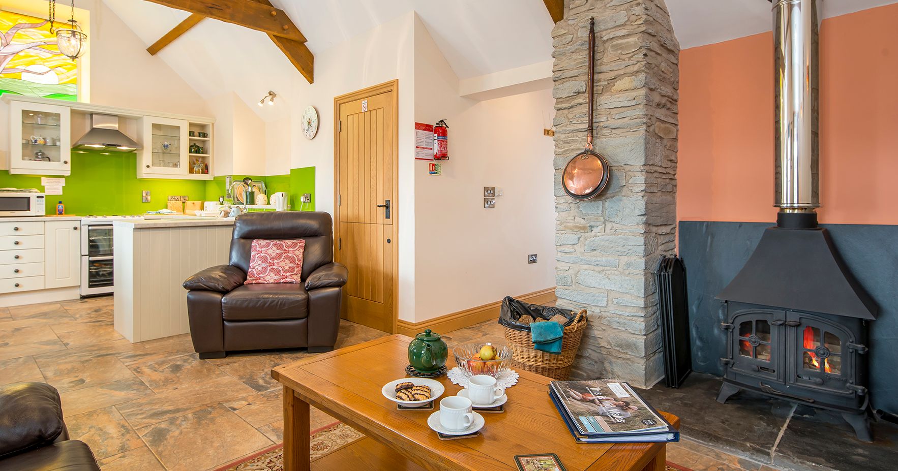 A cozy open-plan living space with a wood-burning stove, a stone feature wall, and a leather armchair. A wooden coffee table is set for tea, with a bright kitchen nearby.