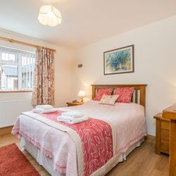 A cozy bedroom in Gwaun Cottage with a wooden bed, pink and white bedding, floral curtains, and warm lighting. A window with blinds overlooks neighboring stone houses.