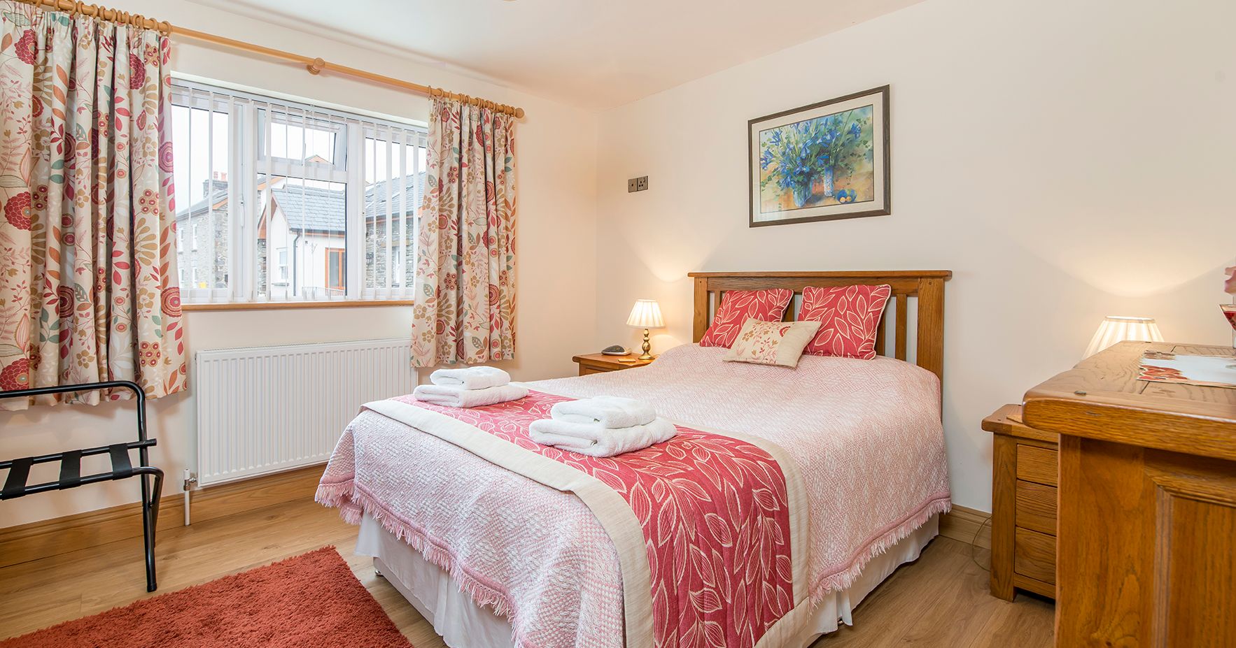 A cozy bedroom in Gwaun Cottage with a wooden bed, pink and white bedding, floral curtains, and warm lighting. A window with blinds overlooks neighboring stone houses.