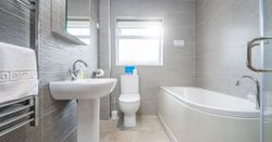 The bathroom in Gwaun Cottage features sleek grey tiles, a white pedestal sink, a curved bathtub, and a toilet beneath a frosted window, with a heated towel rail.