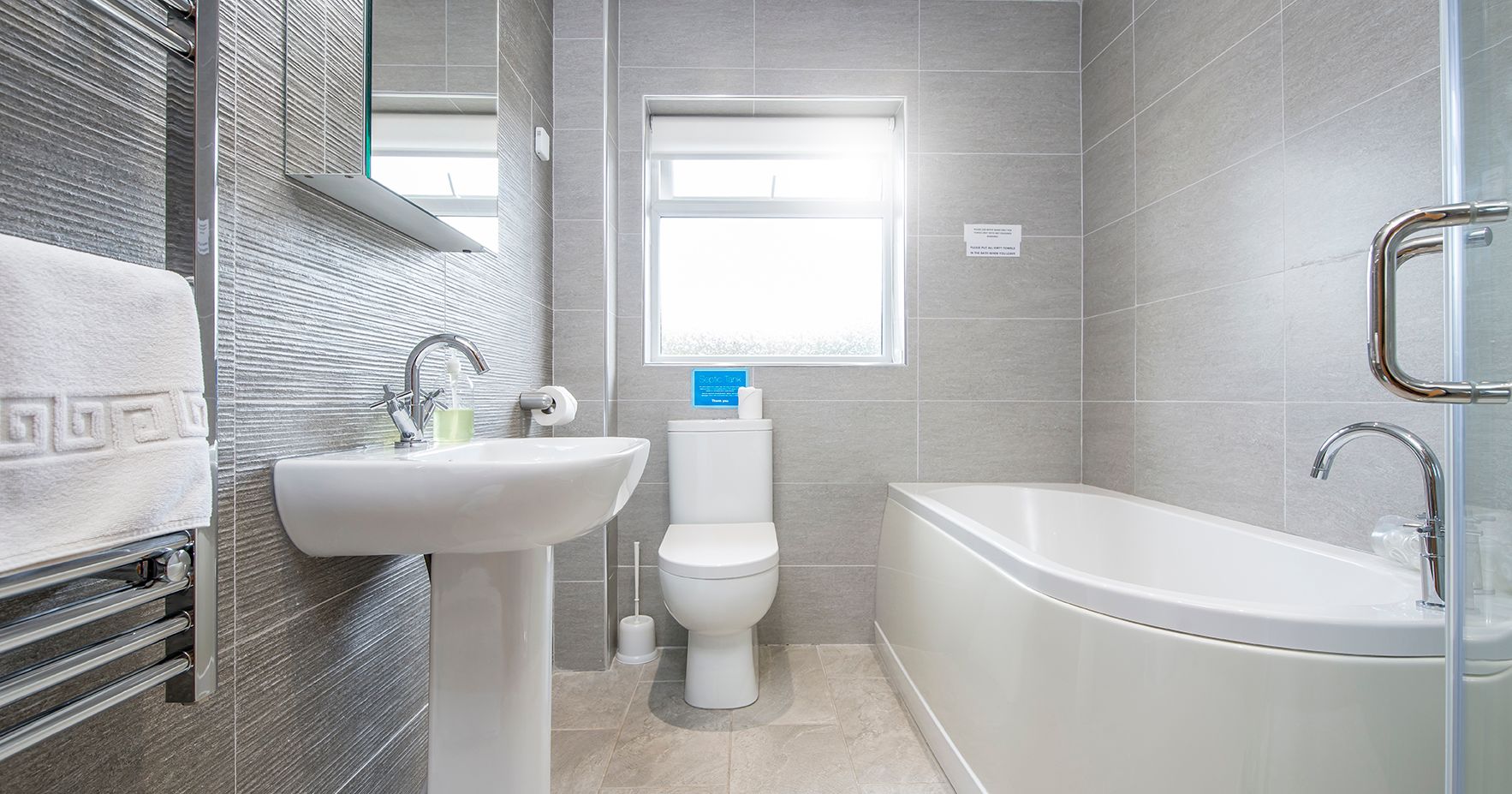 The bathroom in Gwaun Cottage features sleek grey tiles, a white pedestal sink, a curved bathtub, and a toilet beneath a frosted window, with a heated towel rail.