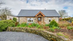 Gwaun Cottage, a charming stone house with a slate roof and wooden porch, is surrounded by a vibrant garden with tulips and greenery, set beside a gravel driveway.