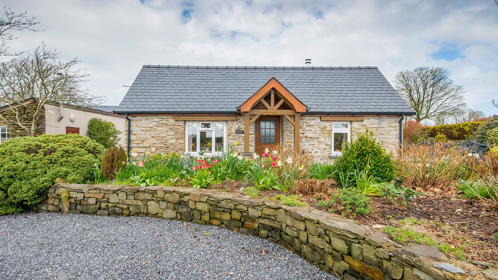 Gwaun Cottage, a charming stone house with a slate roof and wooden porch, is surrounded by a vibrant garden with tulips and greenery, set beside a gravel driveway.
