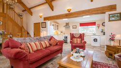 A cozy open-plan living space with a red sofa, armchair, and wooden coffee table set for tea. The room features exposed beams, a staircase, and a bright kitchen in the background.