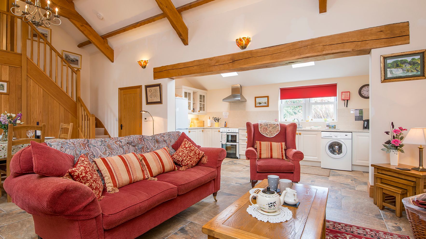 A cozy open-plan living space with a red sofa, armchair, and wooden coffee table set for tea. The room features exposed beams, a staircase, and a bright kitchen in the background.