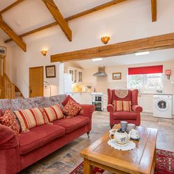 A cozy open-plan living space with a red sofa, armchair, and wooden coffee table set for tea. The room features exposed beams, a staircase, and a bright kitchen in the background.